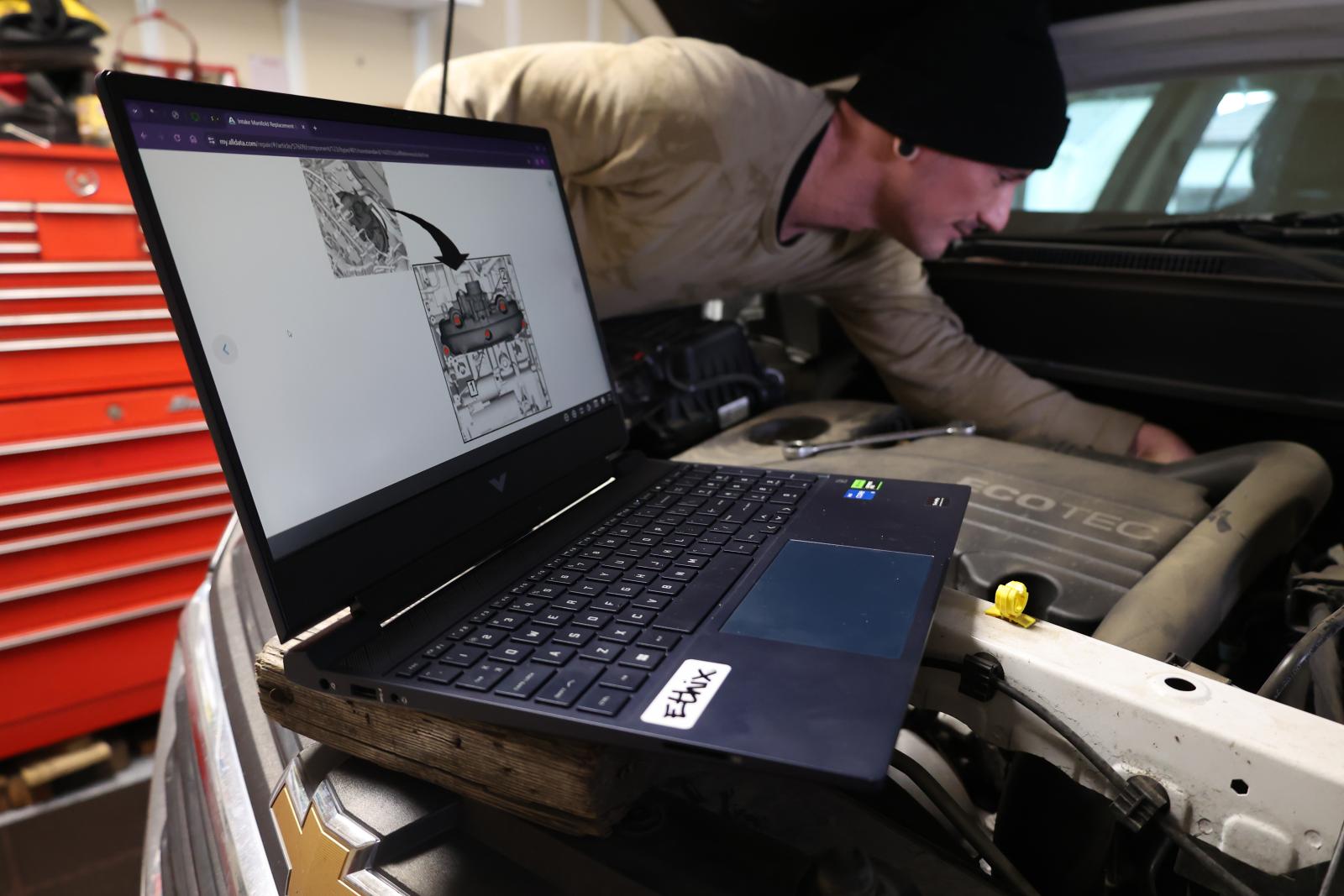 man looking in car engine with alldatadiy repair information on a laptop screen in the foreground